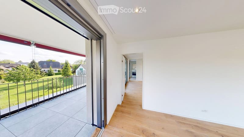 Bright hallway with wooden flooring, white walls, and a large sliding glass door leading to a balcony with a view of a green garden and neighboring houses.