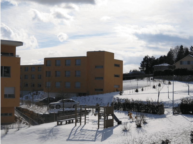 A multi-story building with a yellow facade and numerous windows, surrounded by a snow-covered playground area with benches, swings, and a slide.