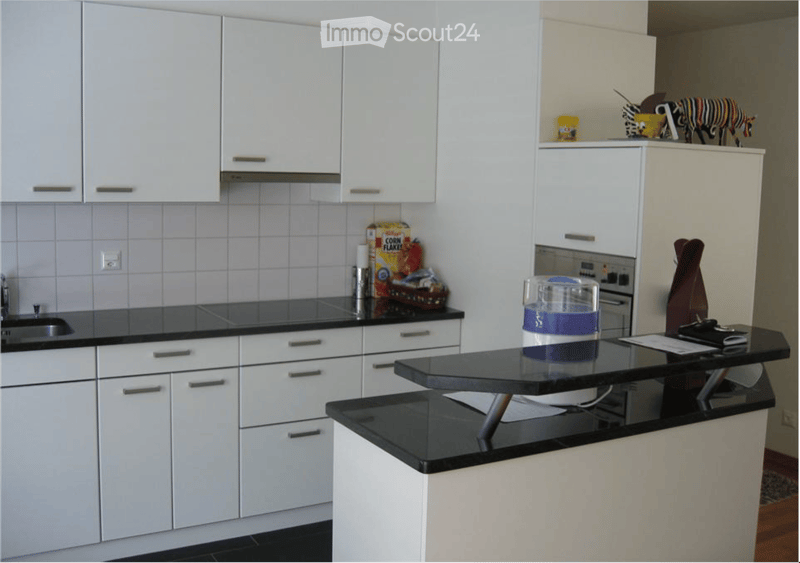 Modern kitchen with white cabinets, black countertops, and stainless steel appliances. Includes a sink, fridge, oven, and a small table with items on it.