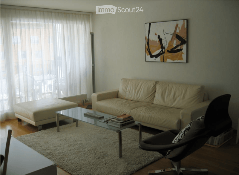Modern living room with a cream-colored sofa, glass coffee table, black leather chair, ottoman, and a large abstract painting on the wall. The room features large windows with white curtains and a shaggy white rug on the wooden floor.