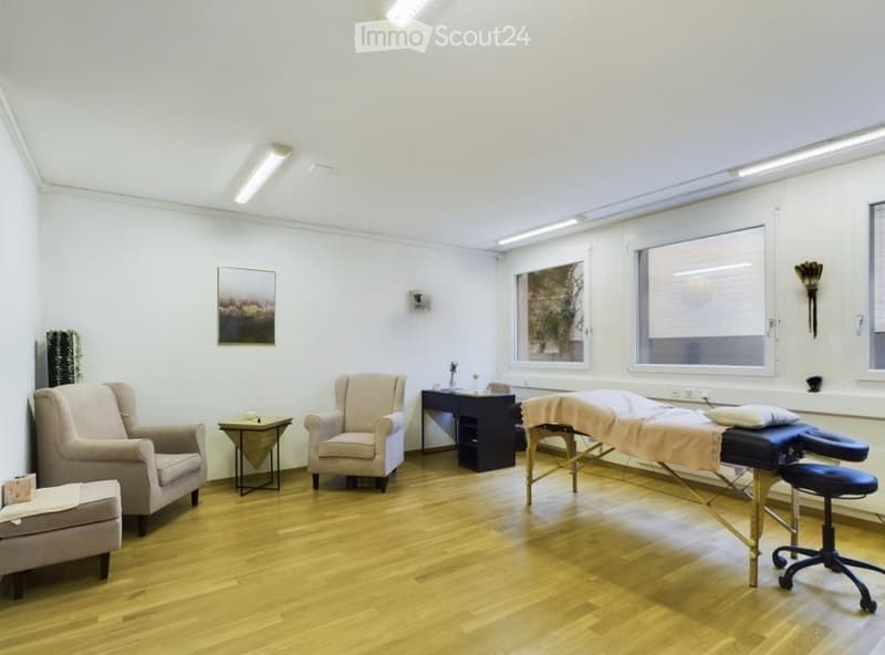 Bright, minimalist room with wooden flooring, two armchairs, a small table, a massage table, and a stool. Large windows provide natural light.