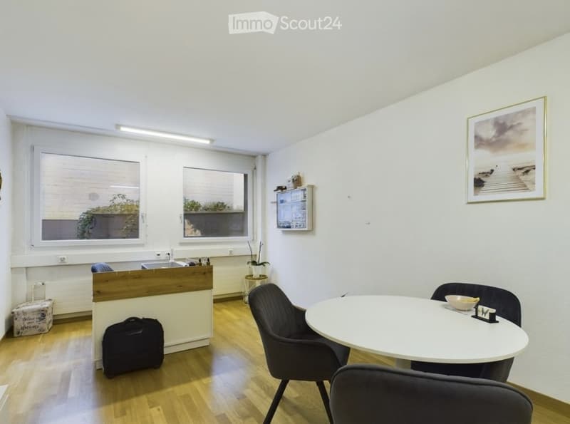 A modern, well-lit room with wooden flooring, white walls, and large windows. Features include a round table with chairs, a sink area, a framed picture on the wall, a small plant, and a suitcase near the wall.