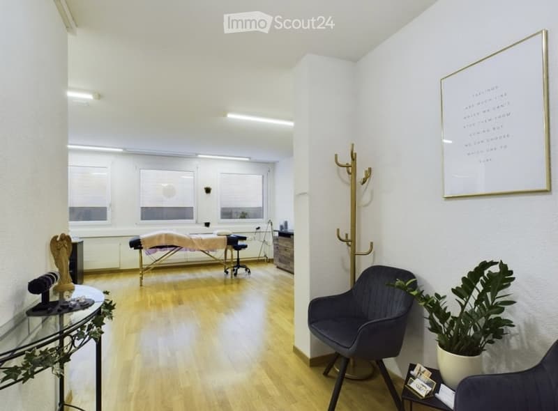 Bright, minimalist room with wooden flooring, large windows, massage table, dark blue chair, small table with plant, framed artwork, and coat rack.