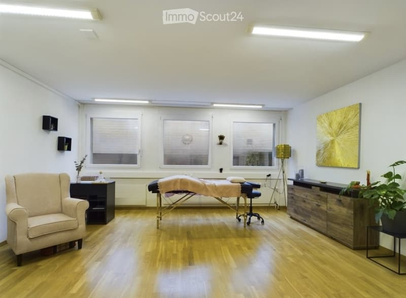 A well-lit, modern room with wooden flooring, large windows, a massage table, a sofa, a cabinet, a lamp, and decorative plants.