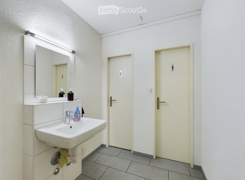 Modern bathroom with a white sink, mirror, and two doors. The sink has a silver faucet and a soap dispenser, with a small cup on the counter. The walls are white, and the floor is tiled.