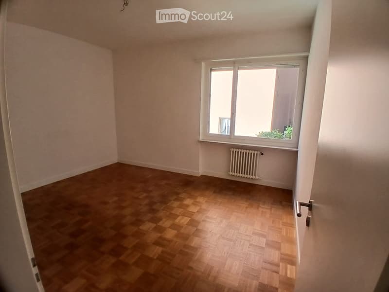 Empty room with wooden flooring, white walls, a window with white frame, and a radiator below the window.