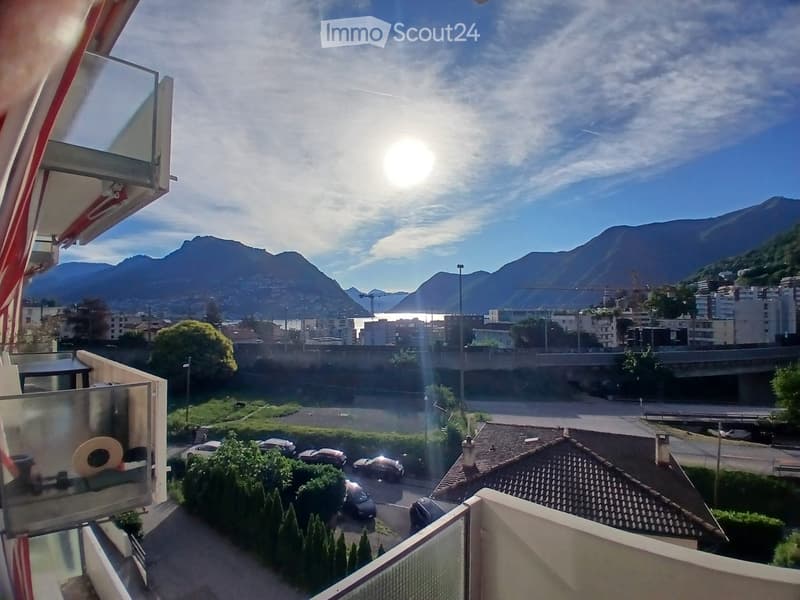 Balcony with a scenic view of a cityscape, mountains, and a body of water in the distance. The foreground shows a parking area with several cars and some greenery.