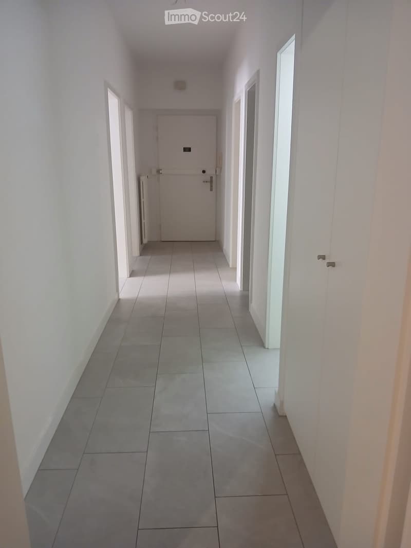 A long, narrow hallway with white walls, tiled floor, and several doors on both sides.