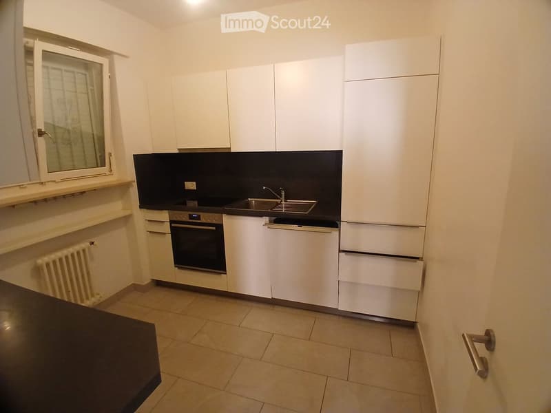 Modern kitchen with white cabinets, a black backsplash, a stainless steel sink, and an oven. The floor is tiled, and there is a radiator on the wall.
