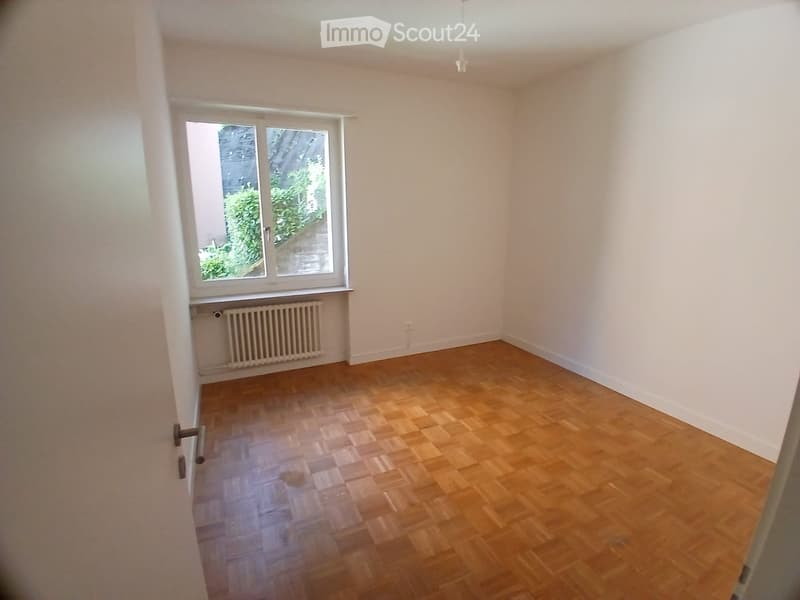 Empty room with wooden flooring, white walls, and a window with a view of greenery. A radiator is visible below the window.