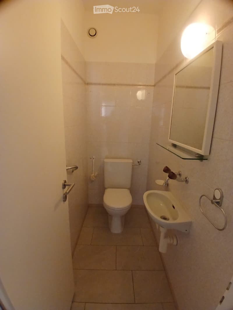 Small bathroom with white tiled walls and floor, featuring a toilet, sink, mirror, and faucet.