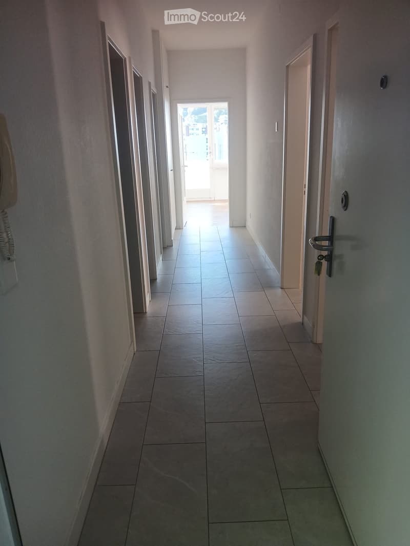 Empty hallway with tiled floor, white walls, and multiple doors leading to rooms. Natural light enters from an open doorway at the end.