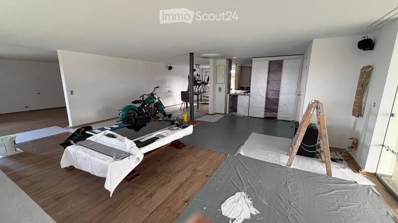 A spacious room with wooden flooring, a table with a laptop, a motorcycle, and several stools. The room appears to be under renovation with protective coverings on the floor and walls.