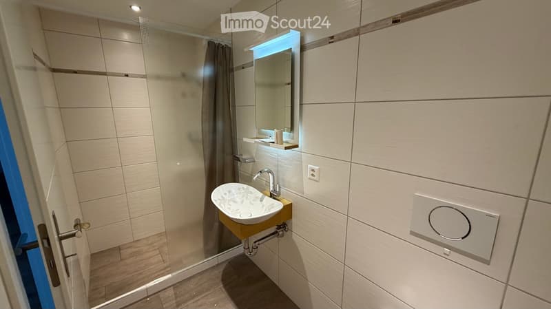 Modern bathroom with a white sink, mirror, and glass shower enclosure. The walls are tiled in light beige with a wooden floor.