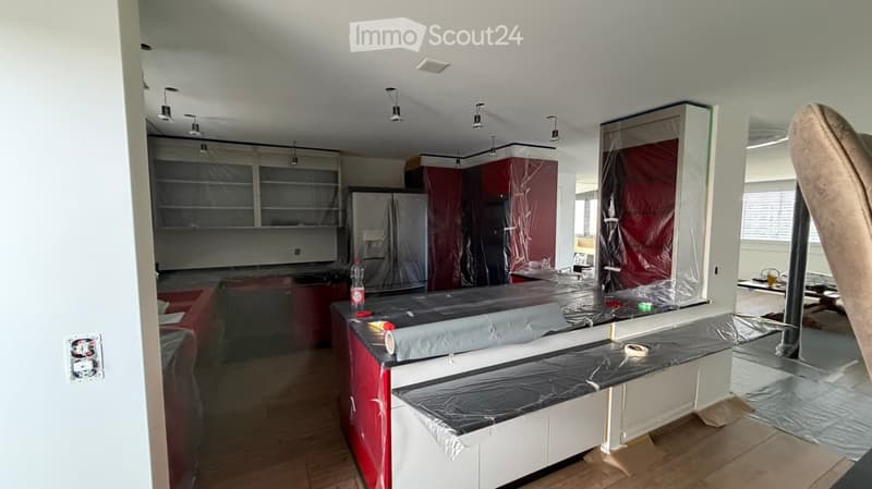Empty kitchen with cabinets covered in protective plastic, countertops covered in black plastic, and a refrigerator in the background.