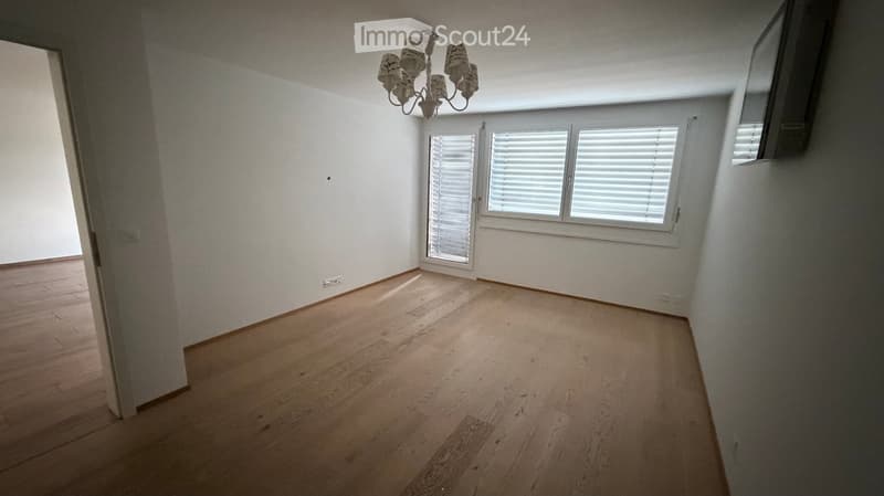 Empty room with wooden flooring, white walls, and large windows with blinds. A chandelier is visible on the ceiling.