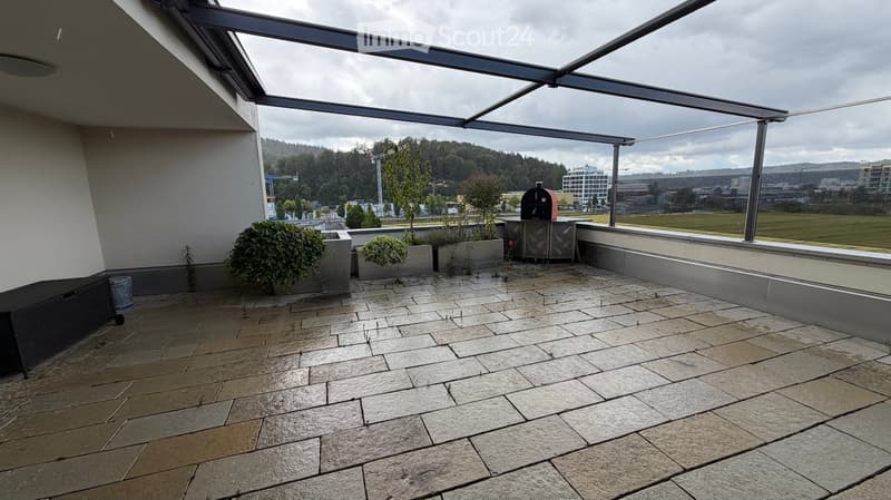 A modern, covered balcony with a tiled floor, potted plants, and a view of a cityscape and greenery.