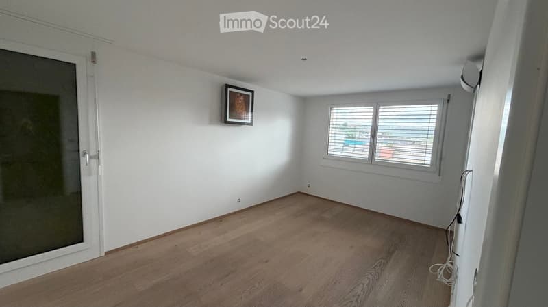 Empty room with wooden flooring, white walls, large window with blinds, and a framed picture on the wall.