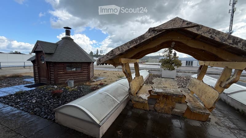 Outdoor seating area with a wooden gazebo, stone table, benches, and a small wooden cabin in the background.