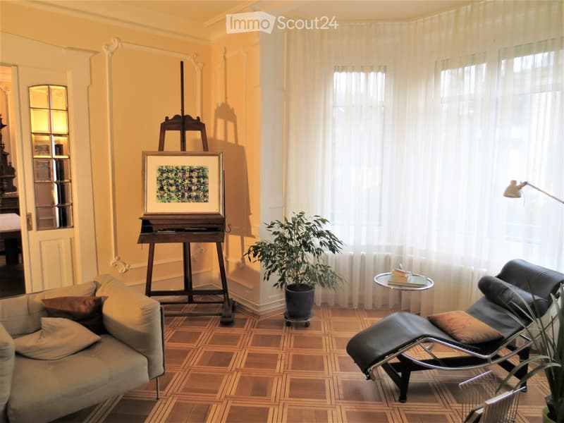 A well-lit living room with a beige sofa, a black leather chair, a small round table with books, a floor lamp, a potted plant, and an easel with a painting.