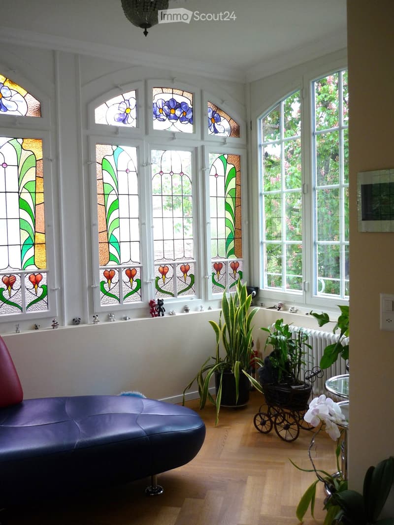 A modern living room with a blue leather sofa, wooden floor, large windows with stained glass, potted plants, and decorative toys on the windowsill.