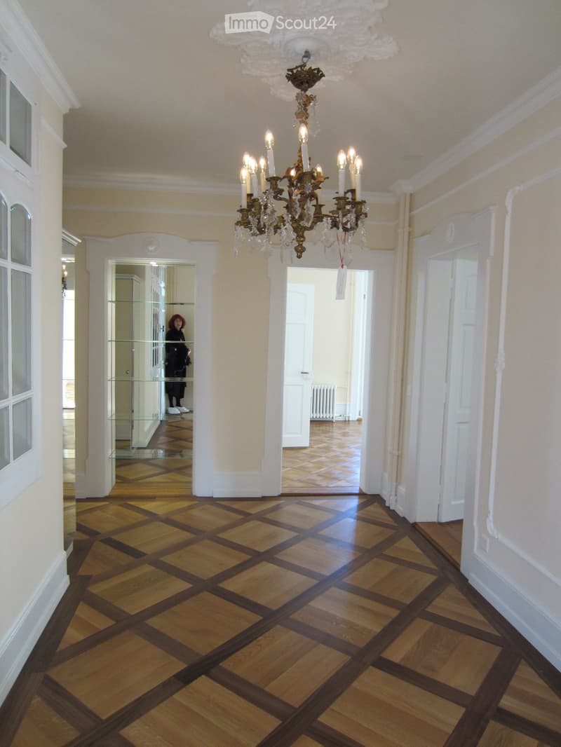 Elegant hallway with a checkered wooden floor, white walls, and a large ornate chandelier with multiple candle-like lights hanging from the ceiling. The hallway features several doors and a glass partition leading to another room.