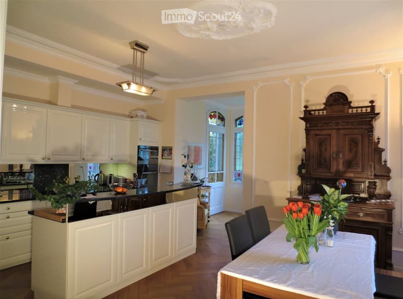 Bright kitchen with white cabinets, black countertop, and a dining area with a table and chairs. Features include a chandelier, pendant light, and decorative wooden cabinet.