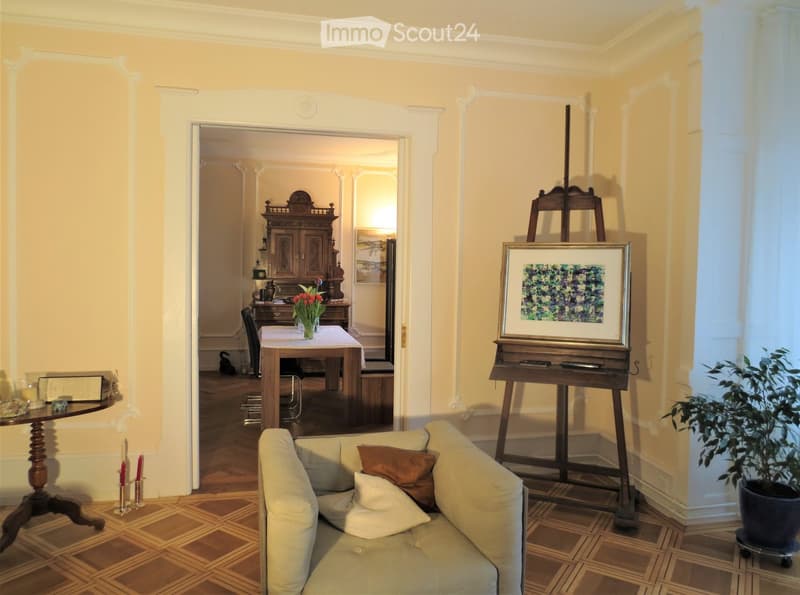 Elegant living room with light yellow walls, wooden parquet flooring, a cream-colored sofa with throw pillows, a small round wooden table with candles, a large easel with a framed painting, and a potted plant near the window. An adjacent room features a w