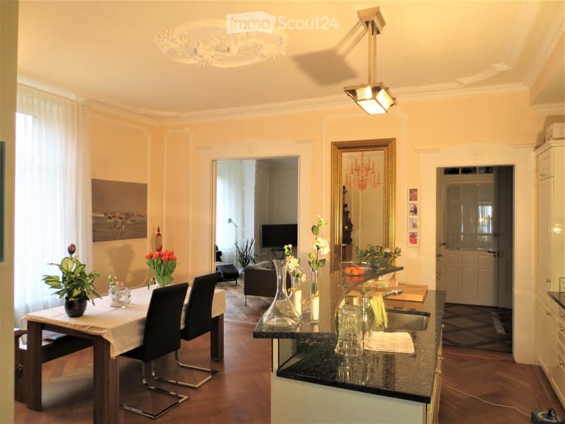 Elegant living and dining area with a dining table, chairs, a modern kitchen counter, vases with flowers, a sofa, and a television in the background.