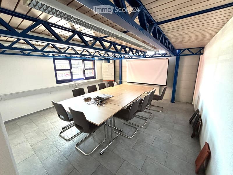 Conference room with a long wooden table, multiple chairs, and a large screen on the wall. The room has a high ceiling with exposed beams and a large window letting in natural light.