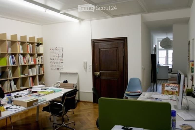 Office space with wooden floor, white walls, bookshelf, desk, chairs, and computer screen.