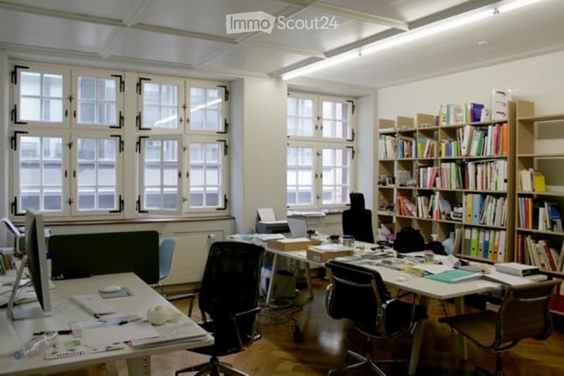 Office space with multiple desks, chairs, computer monitors, keyboards, and bookshelves filled with books.