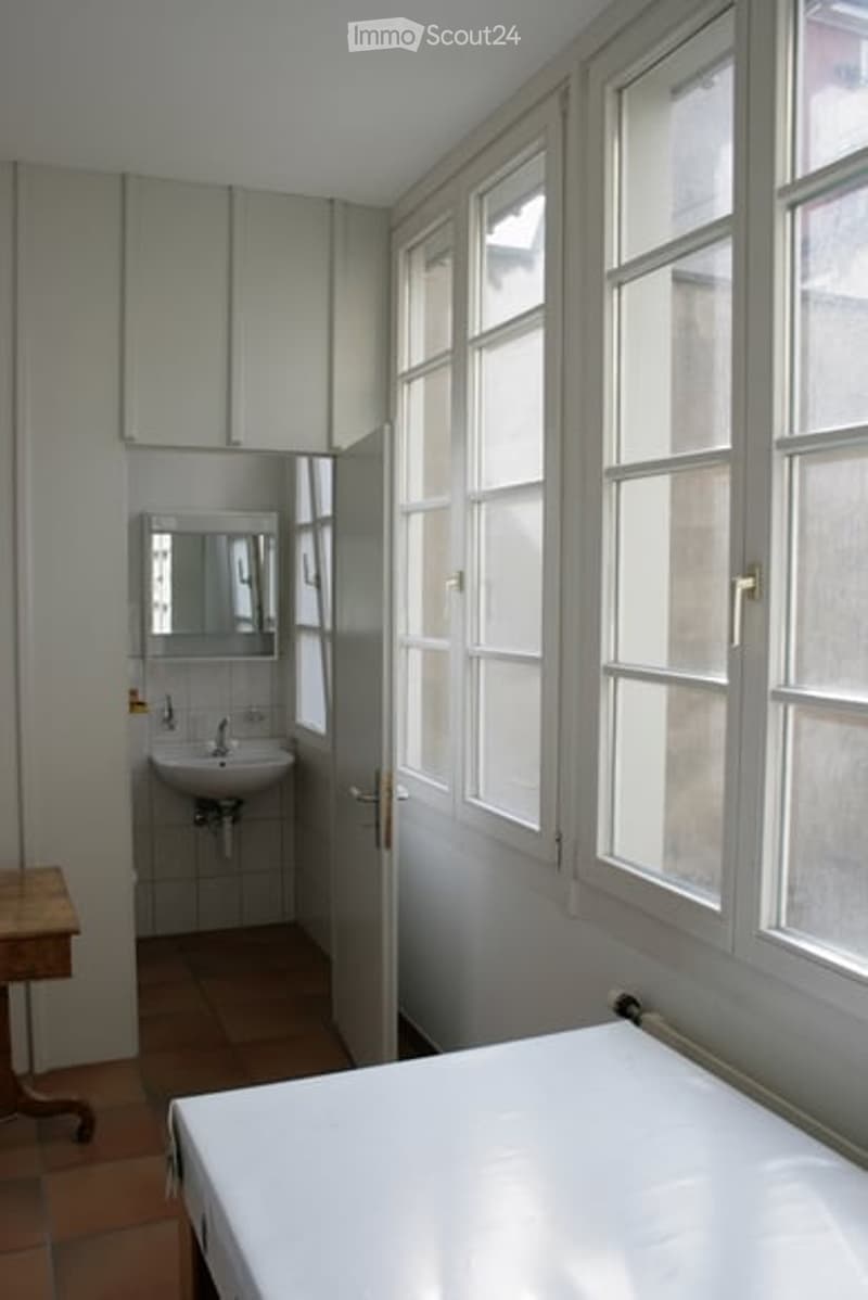 A small, well-lit bedroom with a bed, a sink, and a mirror in an adjacent bathroom area. The room features large windows and a wooden table near the bed.