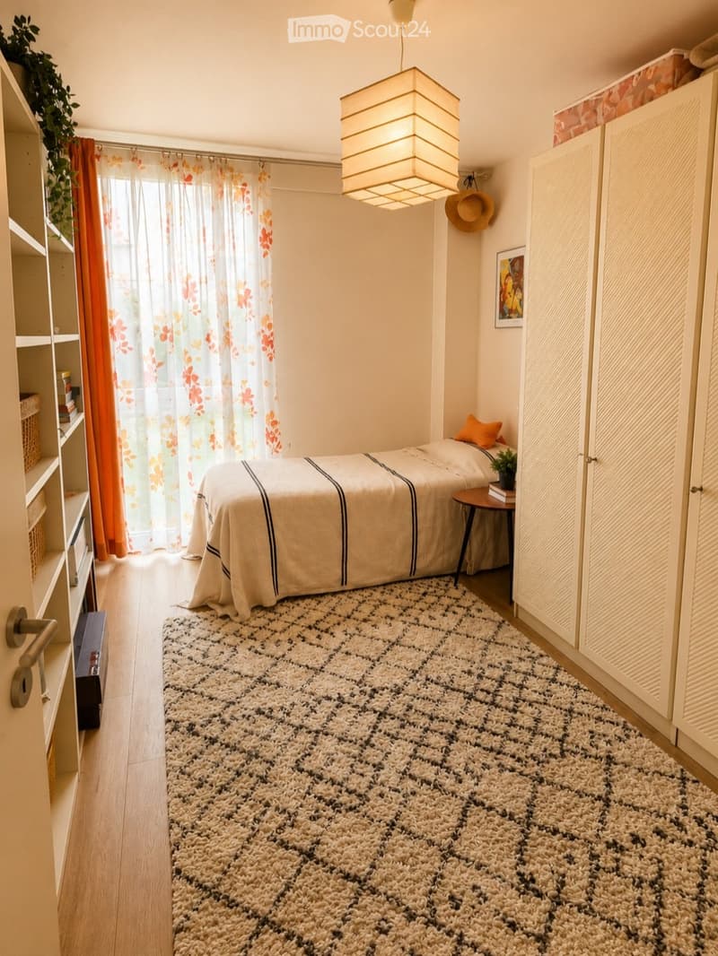 Cozy bedroom with a bed, nightstand, small table, and chair. Features a patterned rug, built-in shelves, and a hanging light fixture.