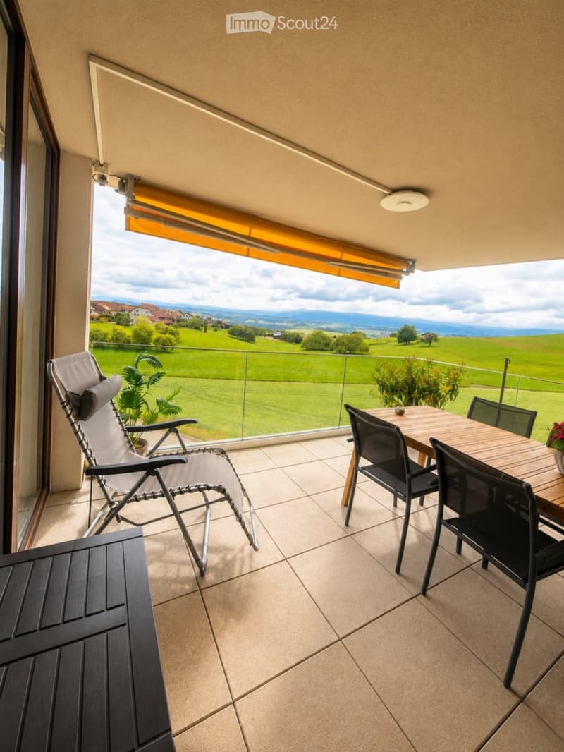 Balcony with tiled floor, glass railing, and scenic countryside view. Includes a wooden table with four black chairs, a folding lounge chair, and potted plants.