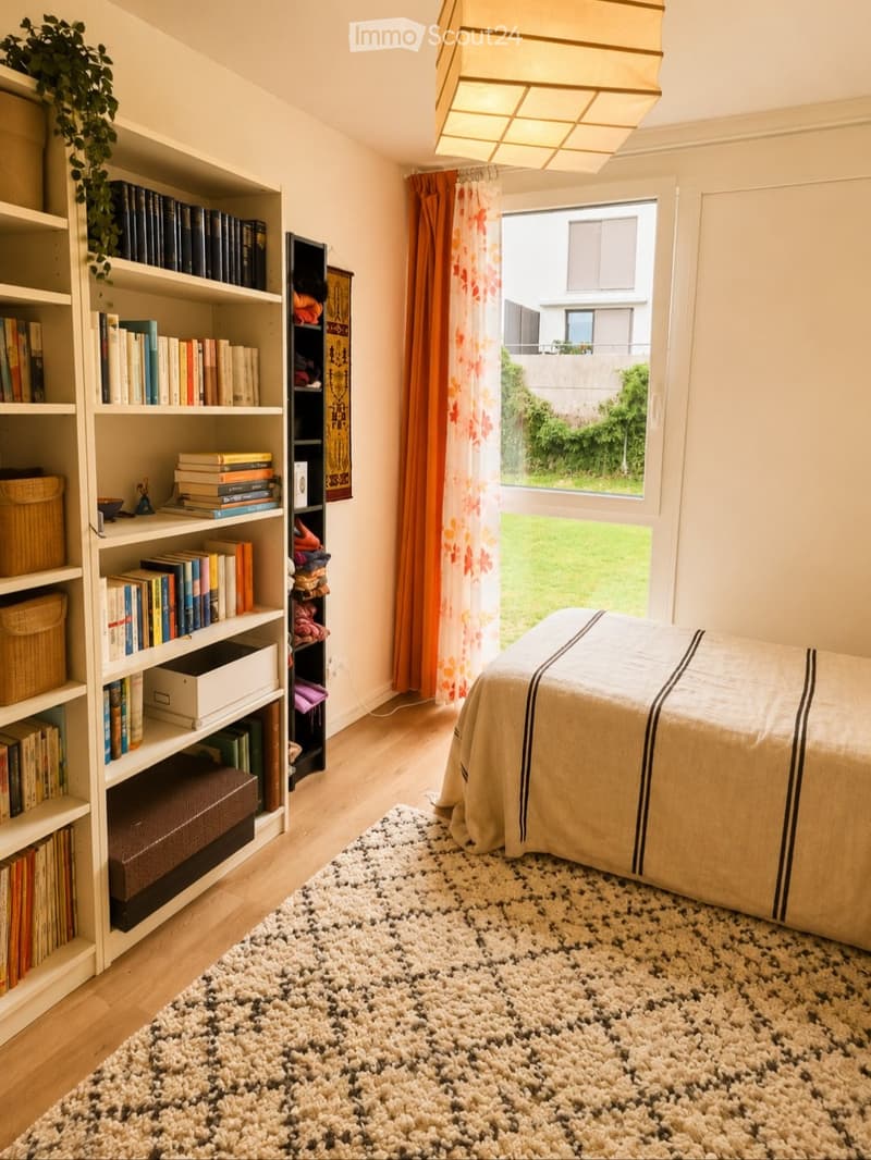 Cozy bedroom with a bed, bookshelf filled with books, a patterned rug, and a window with orange curtains. A hanging lamp provides lighting.