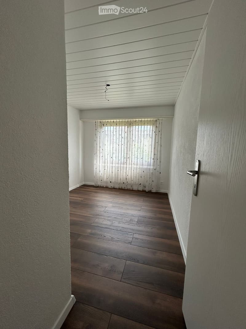 Empty hallway with wooden flooring, white walls, and a window with sheer curtains at the end.