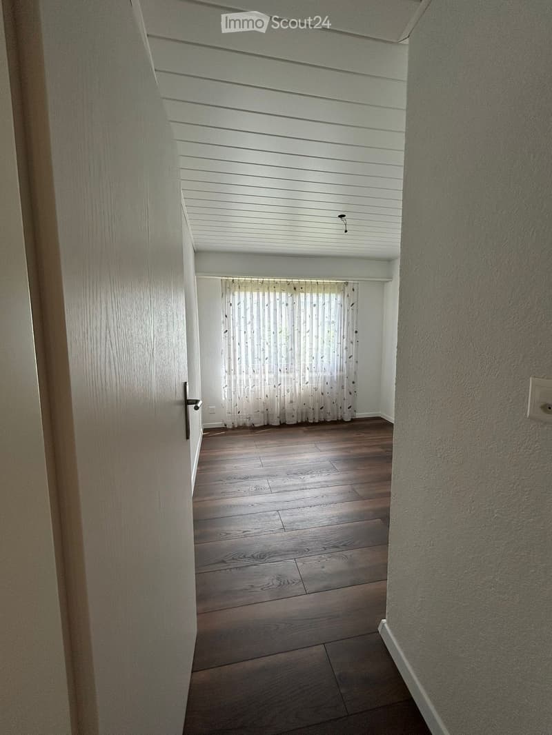 A narrow hallway with wooden flooring, white walls, and a window with sheer curtains at the end.