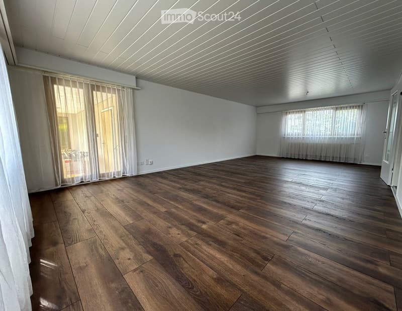 Empty room with wooden flooring, white walls, large windows with sheer curtains, and a white ceiling with linear lighting.