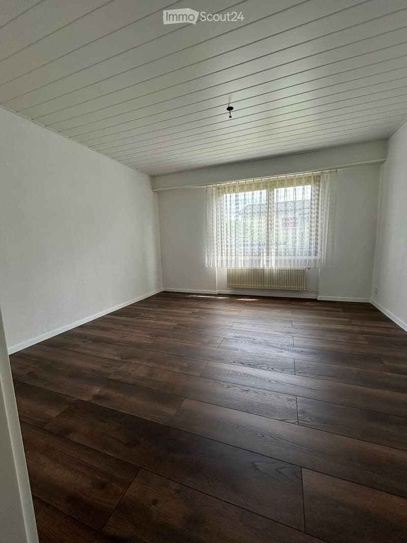 Empty room with wooden flooring, white walls, and a window with sheer curtains. A radiator is visible below the window.