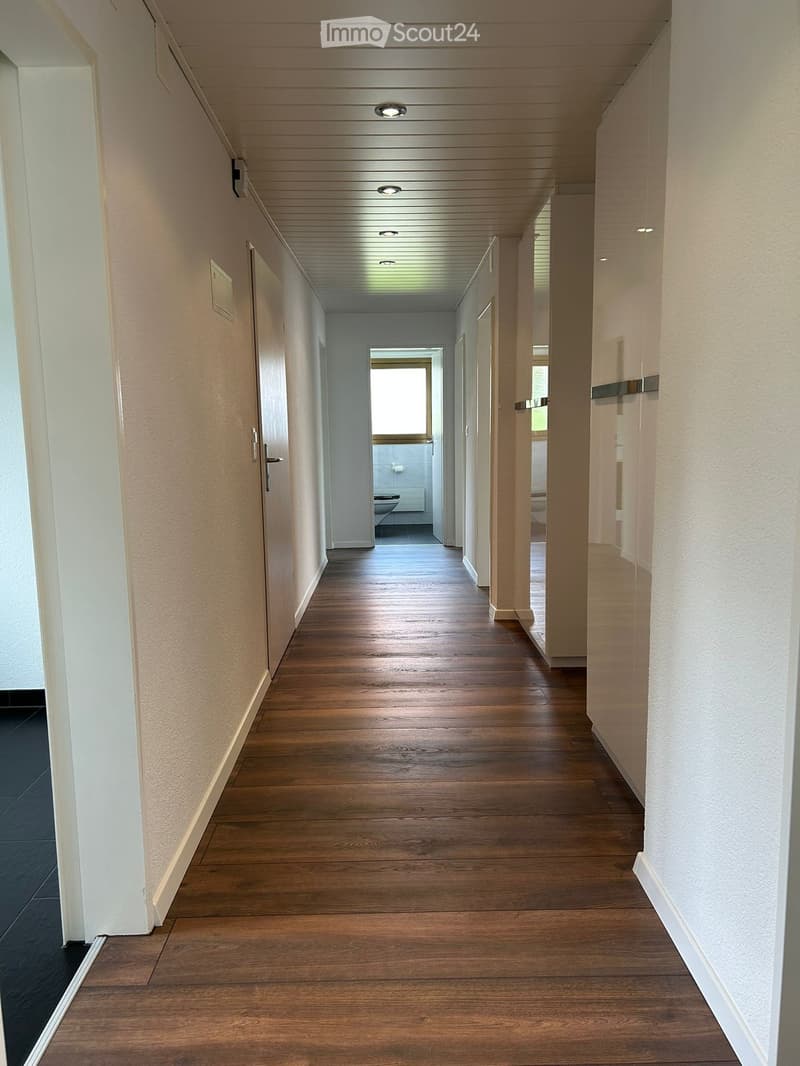 Clean, modern hallway with wooden flooring, white walls, and recessed lighting. Doors lead to rooms, including a visible bathroom at the end.