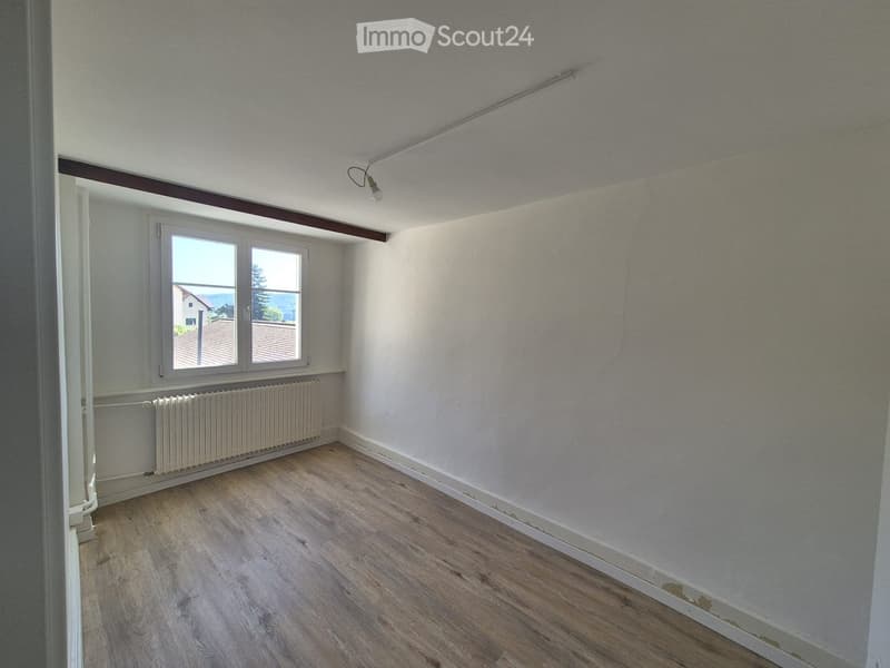 Empty room with light wooden flooring, white walls, a window with a view, and a radiator under the window.
