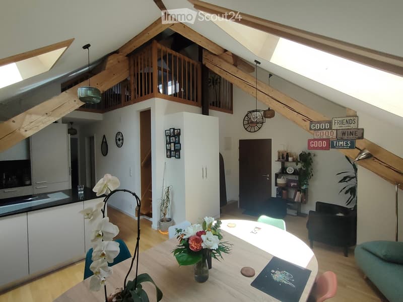 Bright, open living and dining area with exposed wooden beams, modern kitchen, dining table with chairs, sofa, decorative signs, and potted plants.