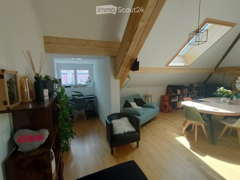 Bright, open-plan living and dining area with wooden floors, exposed wooden beams, skylight windows, a blue sofa, a black armchair, a round dining table with chairs, a bookshelf, and a small piano area.