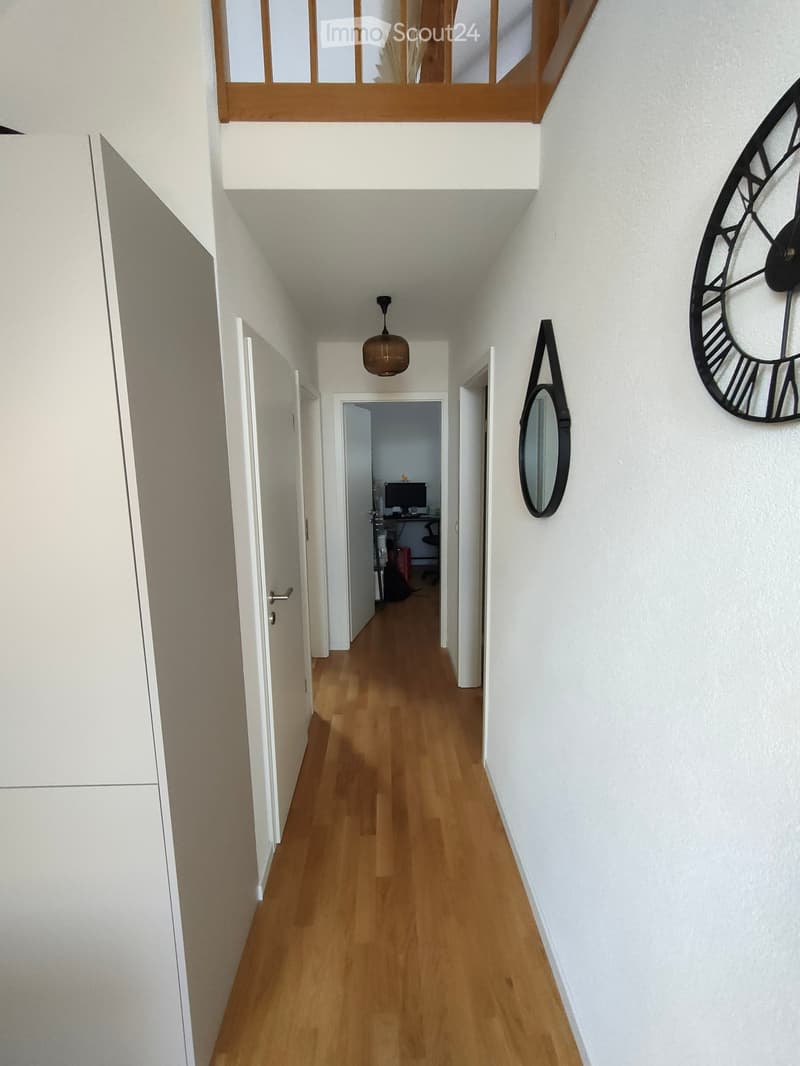 A narrow hallway with wooden flooring, white walls, and several doors leading to different rooms. A large wall clock and a mirror are visible on the right side.