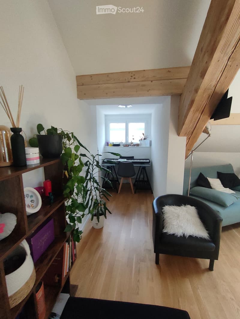 A modern, open-concept living space with wooden flooring, a green sofa, a black armchair, a small table with a keyboard, a wooden shelf with books and decorative items, and a large potted plant.