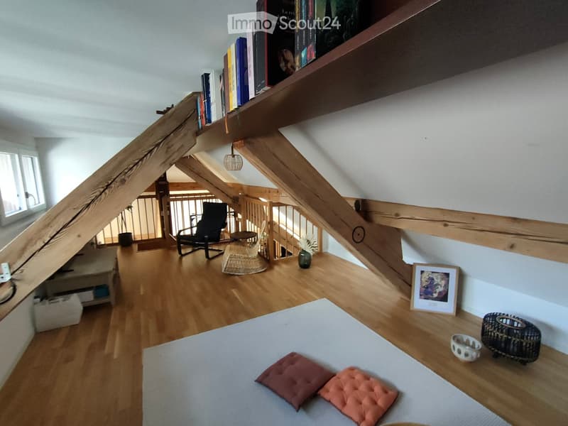 Cozy living space with wooden floors, a staircase leading to another level, a chair, small table, and decorative items including a shelf with books and a framed picture.