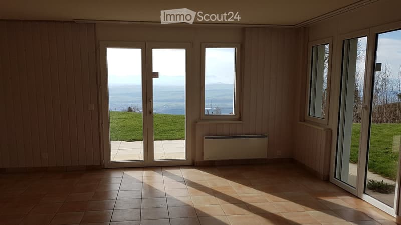 Empty room with tiled floor, wooden walls, large windows, and sliding glass doors opening to a grassy area.