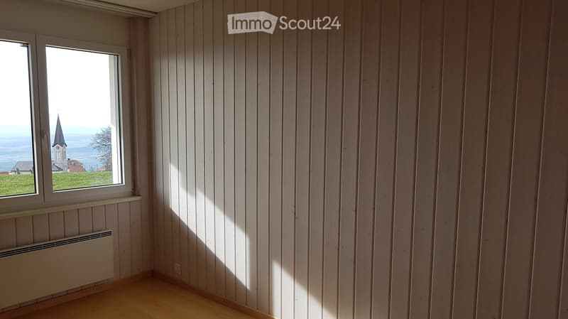 Empty room with wooden panel walls and a wooden floor, featuring a large window with a view of a church and landscape outside.