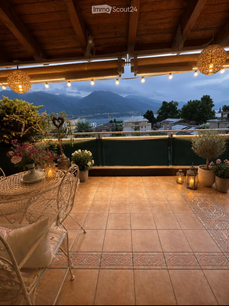 Balcony with terracotta tiles, wooden ceiling, string lights, and a scenic mountain view. Includes a small table, chairs, vases, lanterns, and potted plants.